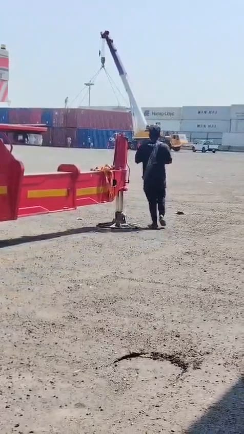 فيديو لكرين متحرك مثبت على أرجله داخل ساحة حاويات ينفذ رفعة، وأمامه تريلة سطحة نازلة على رجل التثبيت ومراقب أرضي يوجّه